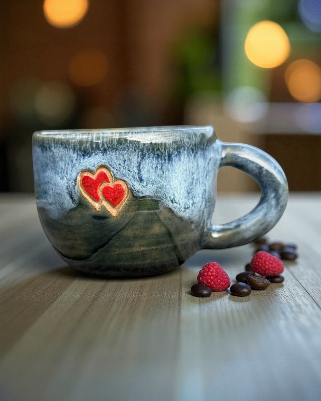 Blue glazed stoneware mug with hearts on a wooden table with coffee beans and raspberries.