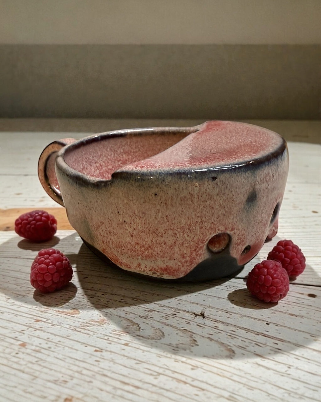 Ceramic berry rinsing mug with soft strawberry glaze beside fresh raspberries on a wooden table.